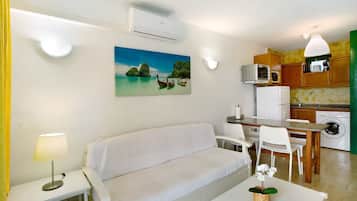 Ett kompakt bostadsutrymme med en soffa, ett matbord, stolar, ett litet köksutrymme med mikrovågsugn och tvättmaskin samt en tavla som föreställer en tropisk scen.