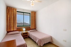 A small room with two single beds, a wooden nightstand, a table, a window with curtains, and a ceiling fan.