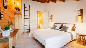 A bedroom with a wooden floor, a bed with white bedding, a wooden headboard, a small stool, a table with a plant, and a ladder against the wall.