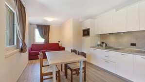 A compact living space with a dining table, chairs, a red sofa, a kitchen area with cabinets and appliances, and a window with curtains. - Apartment 'Al Baitin Trepalle' with Mountain View (Livigno)