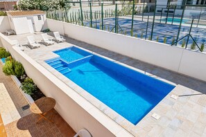 A rectangular swimming pool with a tiled deck, surrounded by a white wall and a green fence. There are lounge chairs and a small table near the pool.