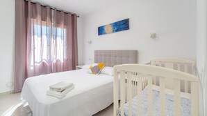 A bedroom with a bed, a crib, a nightstand, a painting, and a window with curtains. - Apartment 'Las Calas Deluxe B' with Shared Pool, Private Terrace and Wi-Fi (Torrevieja)