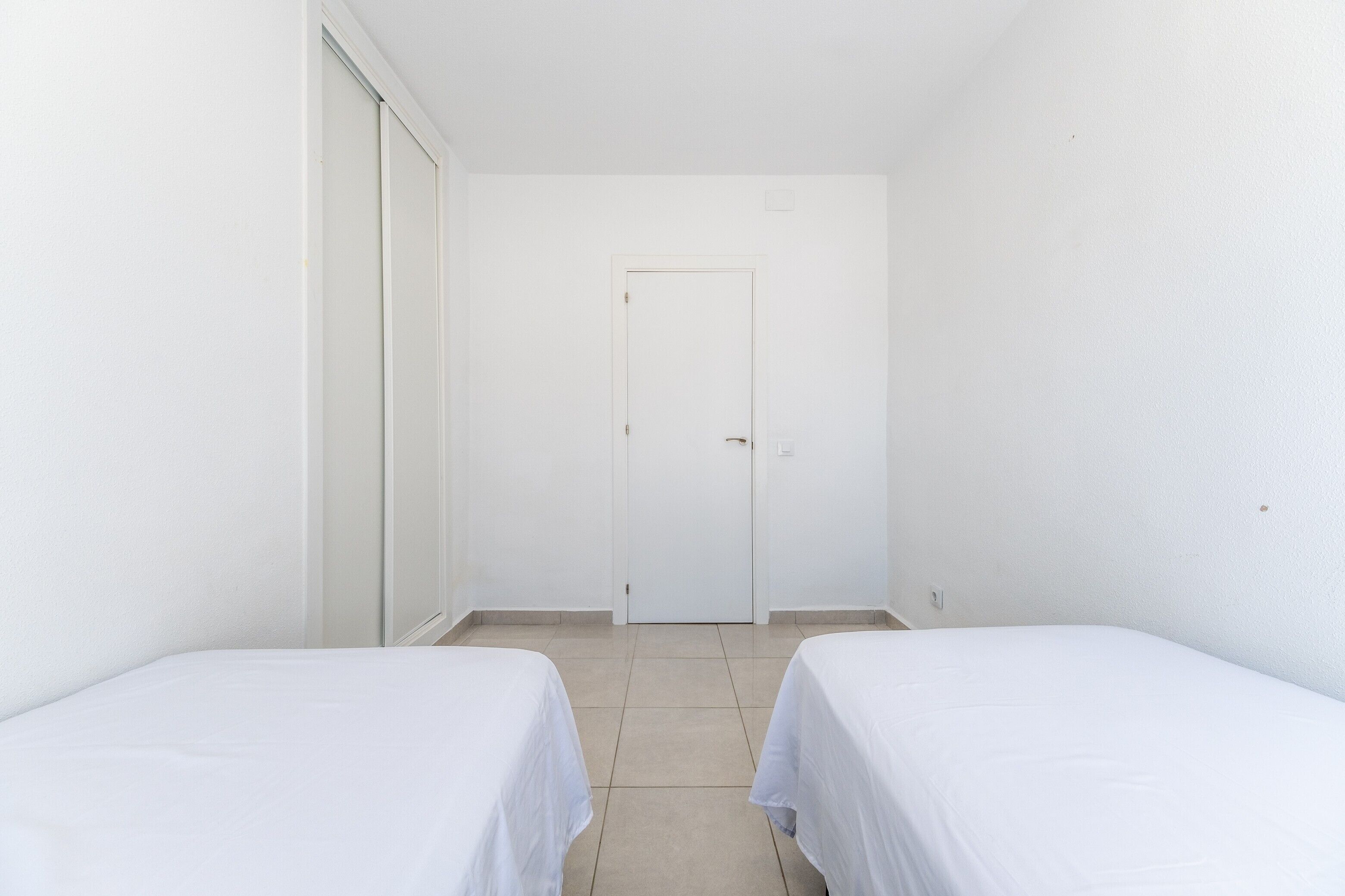 Una habitación con dos camas individuales, una puerta blanca y suelo de baldosas.