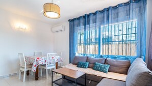 A living room with a sofa, a dining table with chairs, a coffee table, a hanging light fixture, and a window with blue curtains. - Apartment 'Las Calas Deluxe B' with Shared Pool, Private Terrace and Wi-Fi (Torrevieja)