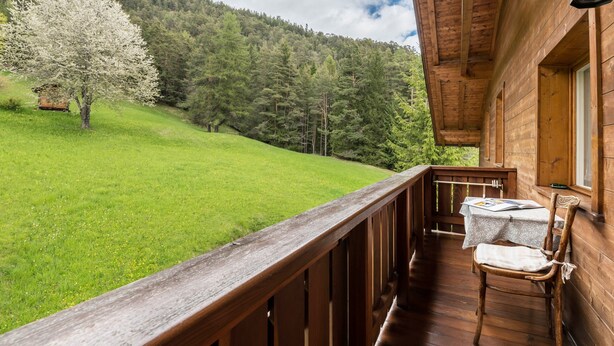 Una cabaña de madera con balcón, una mesa y una silla, y vista a un campo cubierto de hierba y una colina boscosa.