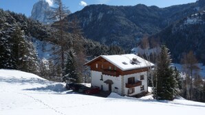 Un refugio de montaña cubierto de nieve con fachada de madera, rodeado de árboles y montañas cargadas de nieve.