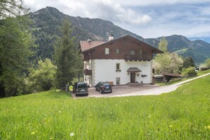 A two-story house with a balcony, parked cars, and a mountain backdrop.
