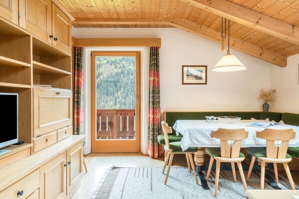 A room with wooden cabinets, a television, a dining table with chairs, a green sofa, a window with a view of trees, and a hanging light fixture.
