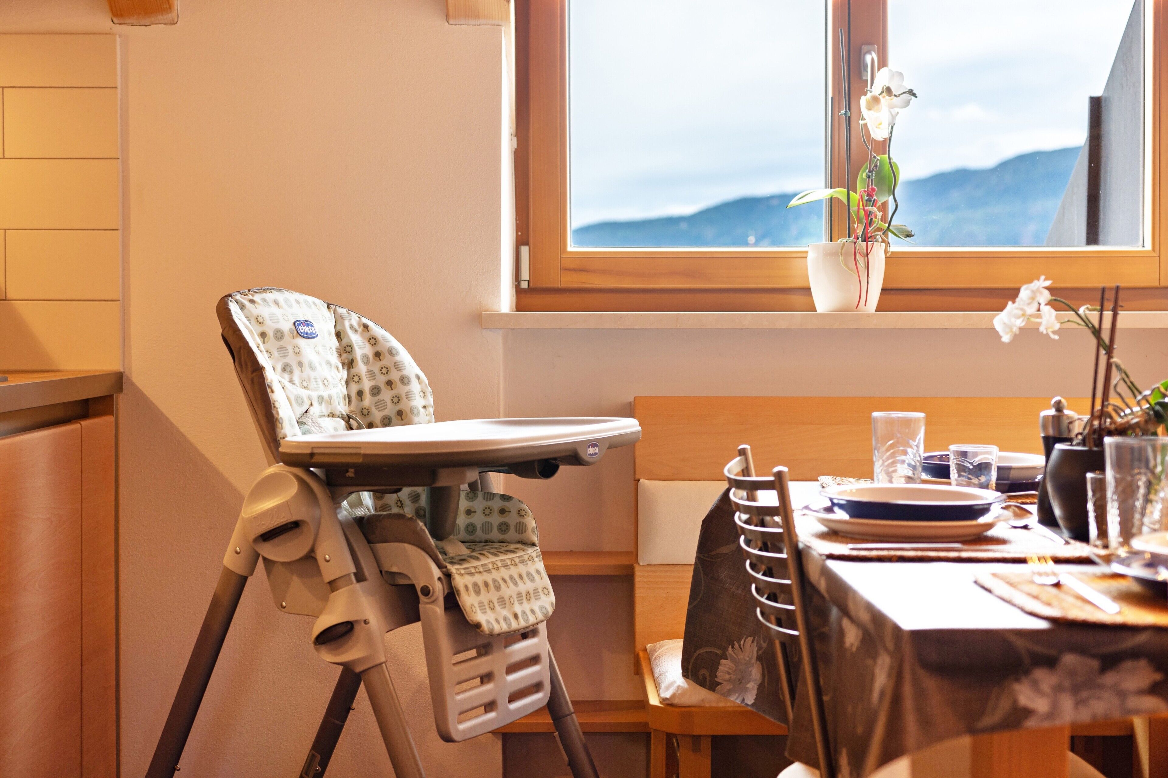 Una silla alta con mesita, una mesa de comedor con platos y vasos, y una ventana con vistas a montañas.