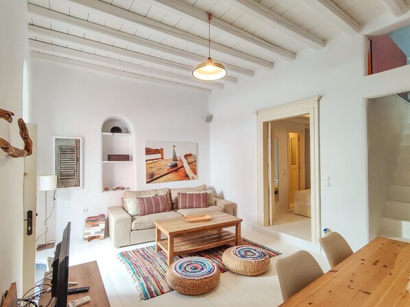 A spacious living area with a wooden coffee table, a sofa, and a dining table. There is a television on a stand, a bookshelf, and a painting on the wall.