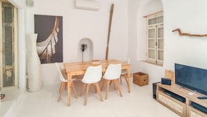 Un coin repas avec une table en bois et des chaises blanches, un téléviseur posé sur un meuble en bois, et une fenêtre donnant sur un paysage agréable.