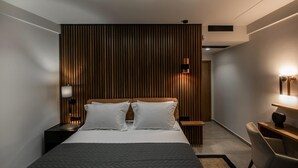 A modern hotel room with a large bed, a wooden accent wall, a nightstand with a lamp, a desk with a chair, and a wall-mounted light fixture.