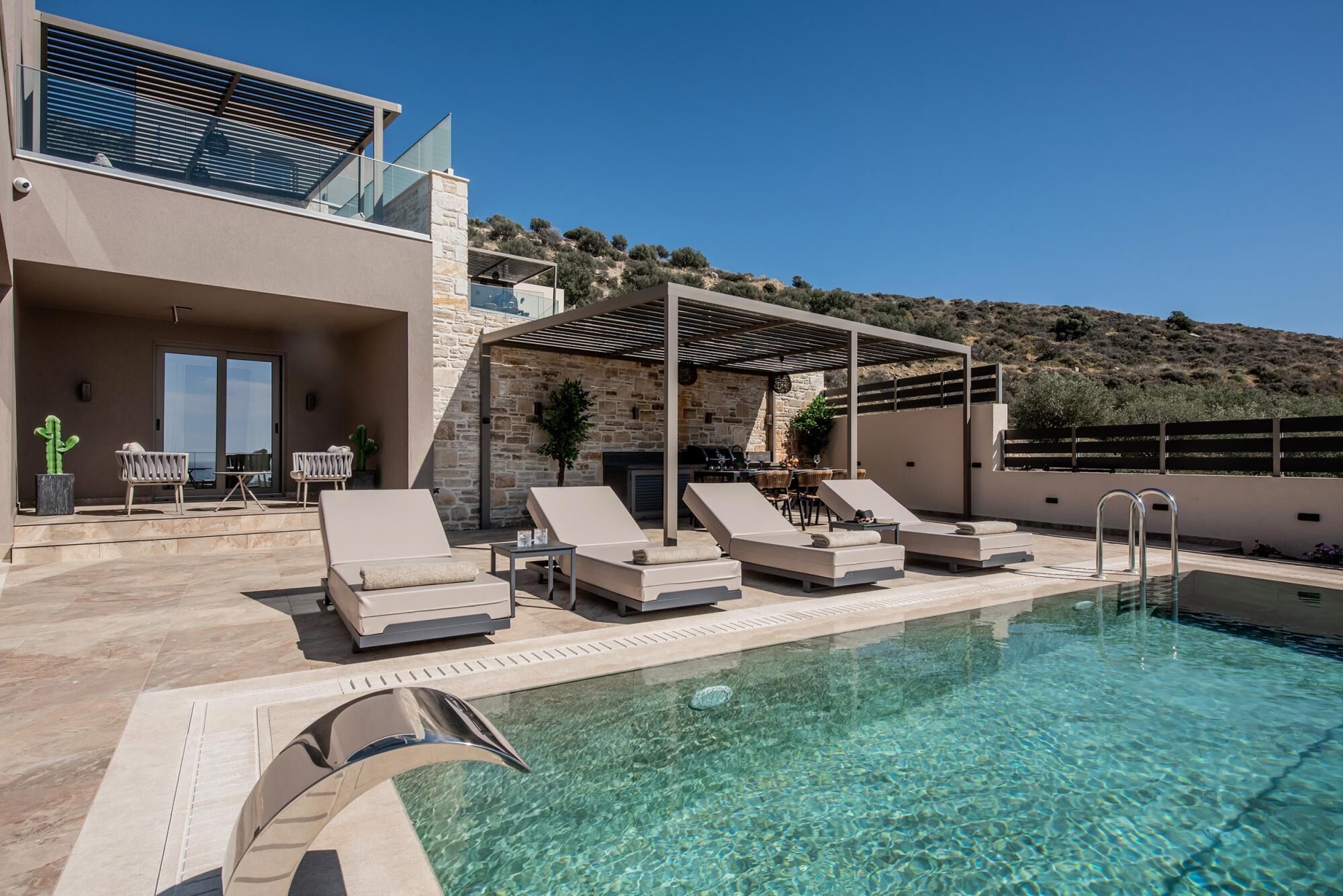 A poolside area with lounge chairs, a barbecue, and a view of the hillside.