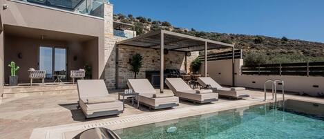 A poolside area with lounge chairs, a barbecue, and a view of the hillside.
