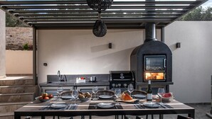 An outdoor kitchen with a pizza oven, grill, and dining table set for a meal. - Villa Paradise 2 in Kalamaki (Faistos)