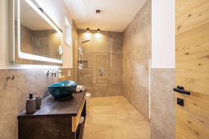 A modern bathroom with a large mirror, a blue vessel sink, a wooden door, and a shower area.