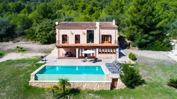 Une maison en pierre à deux étages, dotée d’une piscine, d’un espace extérieur aménagé et offrant une vue sur une colline boisée.