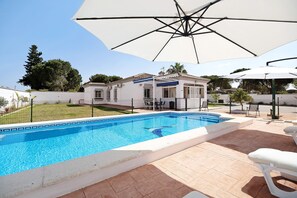 Une piscine bordée de carreaux, un patio avec un parasol blanc et un bâtiment doté d’un toit en tuiles.