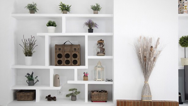 Una pared blanca con varias repisas que sostienen objetos decorativos, como plantas, un farol y una maleta antigua.