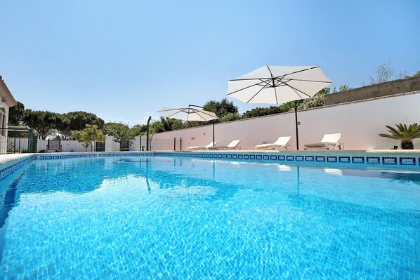 A clear swimming pool with lounge chairs and umbrellas, surrounded by a low wall and greenery.