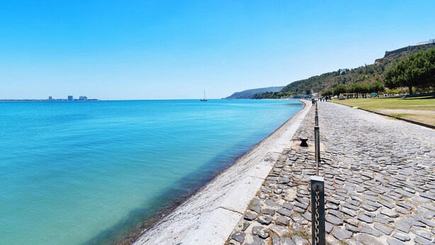 A coastal promenade with a stone pathway, clear blue water, and distant cityscape. - Villa 'Little Paradise 25 Min From Lisbon' with Private Pool, Private Terrace and Wi-Fi (Azeitão)