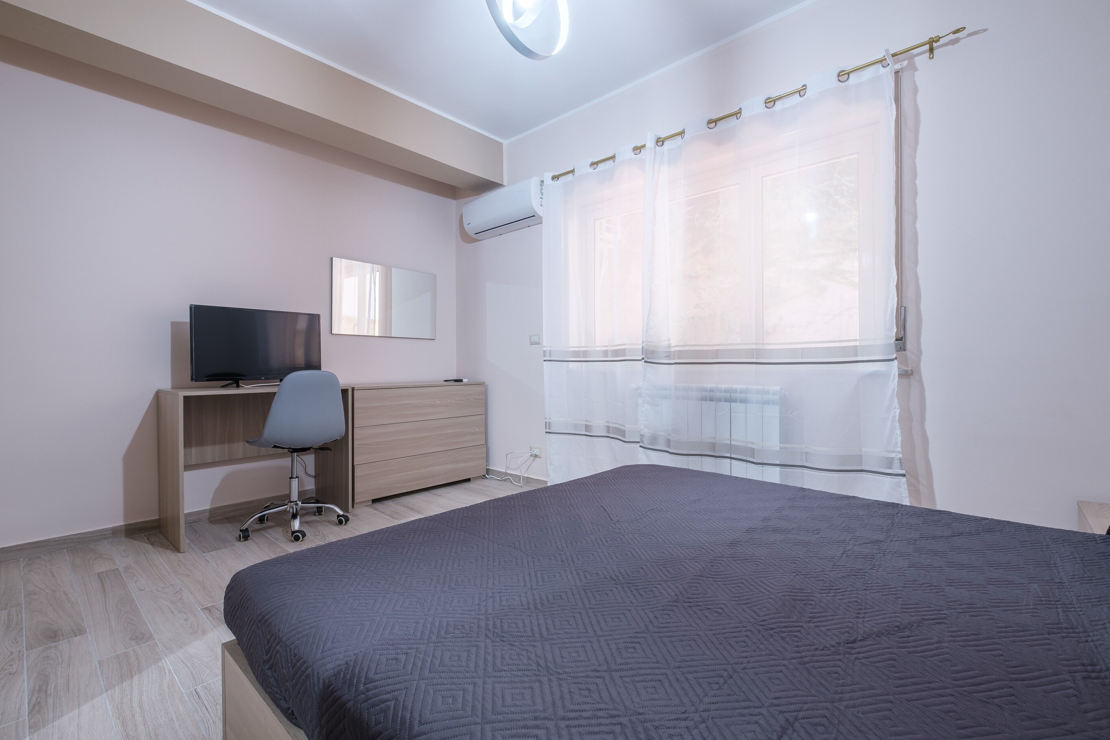 A bedroom with a bed, a desk with a computer, a chair, a dresser, and a window with curtains.