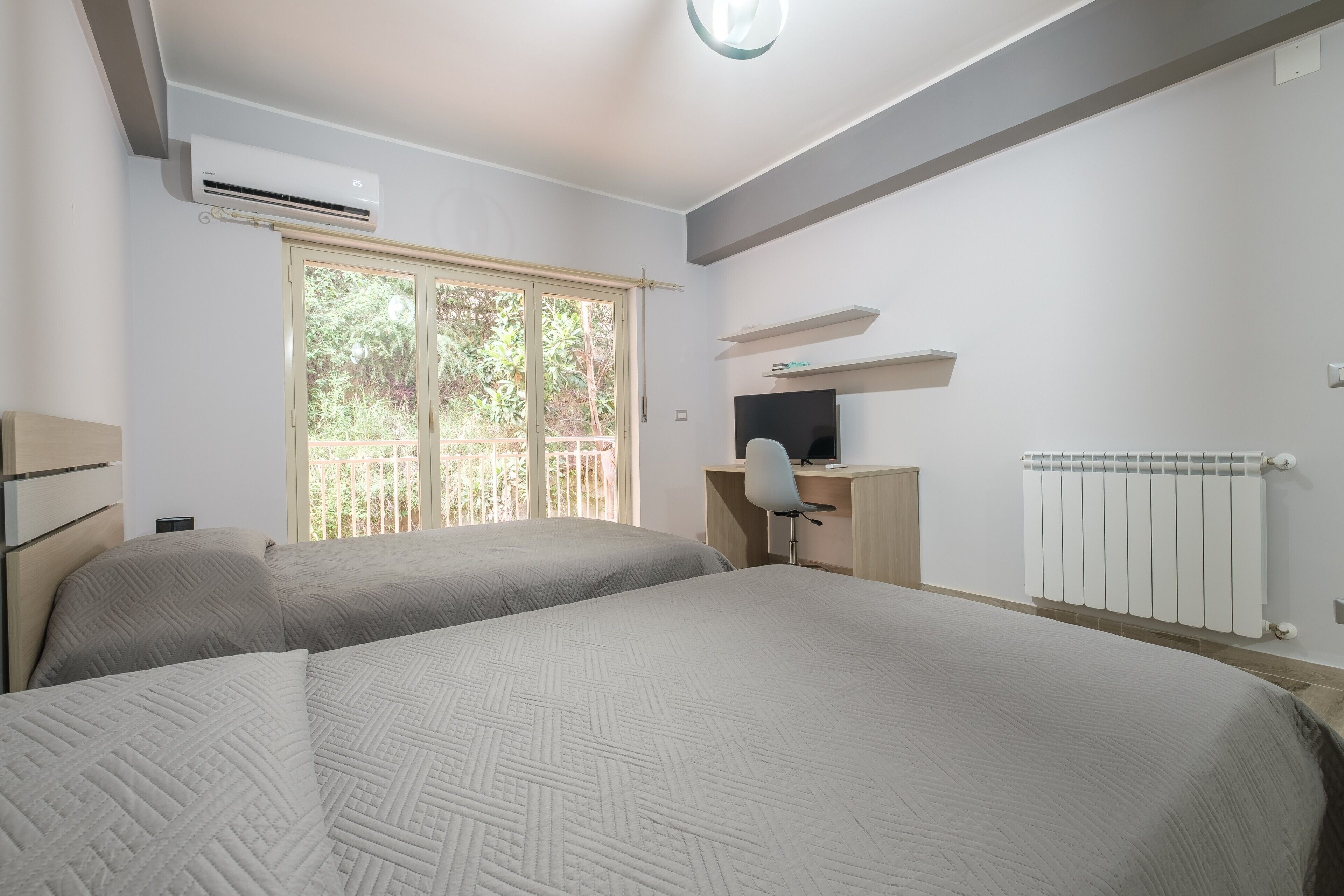 A bedroom with two beds, a desk, a chair, an air conditioner, and a window with a view of greenery.