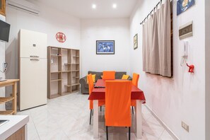 A modern kitchen with a refrigerator, a dining table with orange chairs, a grey sofa, and a wooden bookshelf.
