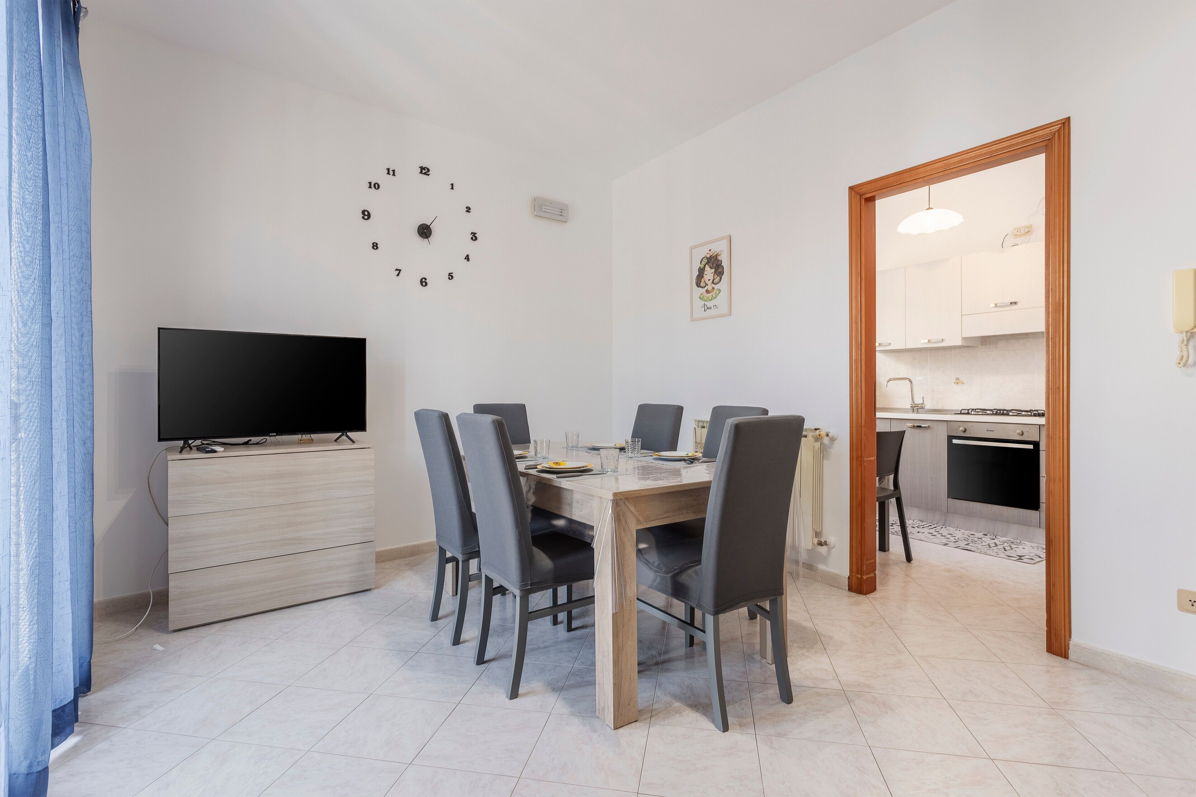 A dining area with a TV, wooden table, grey chairs, and a clock on the wall.