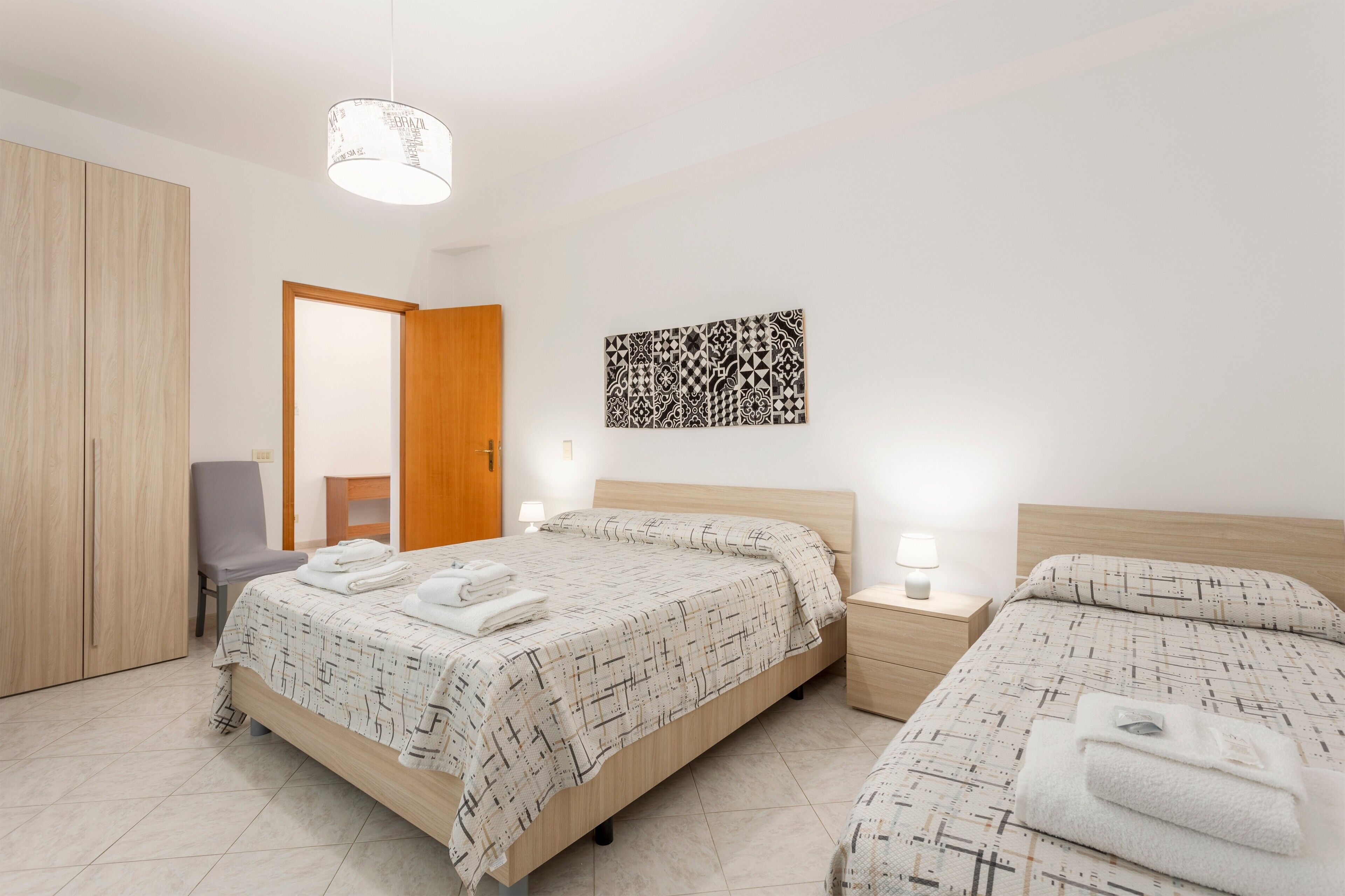A bedroom with two beds, a wardrobe, a chair, a bedside table with a lamp, and a wall art piece.
