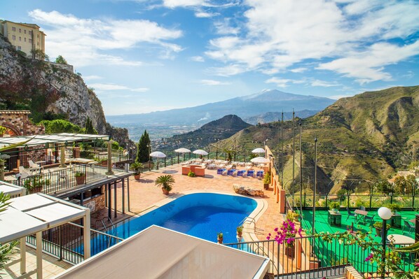 Ein Poolbereich mit Liegestühlen und Sonnenschirmen, Blick auf die Berge und ein Gebäude auf einer Klippe.