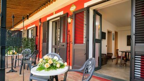 Eine Terrasse mit Sitzgelegenheit im Freien, einem Tisch mit Blumen und einem Gebäudeeingang mit Fensterläden.