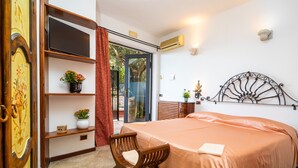 A bedroom with a bed, a wooden chair, a TV, a shelf with a plant, and a door. - Holiday Apartment 'Garden Cottage I Dammusi - Ulivo' with Mountain View, Shared Pool & Wi-Fi (Castelmola)