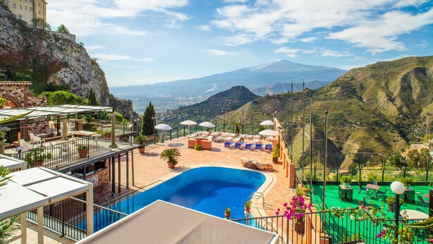 A pool area with lounge chairs and umbrellas, a view of the mountains, and a building on a cliff. - Holiday Apartment 'Garden Cottage I Dammusi - Ulivo' with Mountain View, Shared Pool & Wi-Fi (Castelmola)