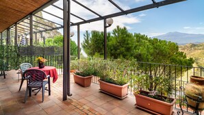 A covered outdoor area with a table and chairs, potted plants, and a view of a mountainous landscape. - Holiday Apartment 'Garden Cottage I Dammusi - Ulivo' with Mountain View, Shared Pool & Wi-Fi (Castelmola)