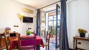 A dining area with a table set for four, a television mounted on the wall, and a sliding glass door leading to an outdoor patio. - Holiday Apartment 'Garden Cottage I Dammusi - Ulivo' with Mountain View, Shared Pool & Wi-Fi (Castelmola)