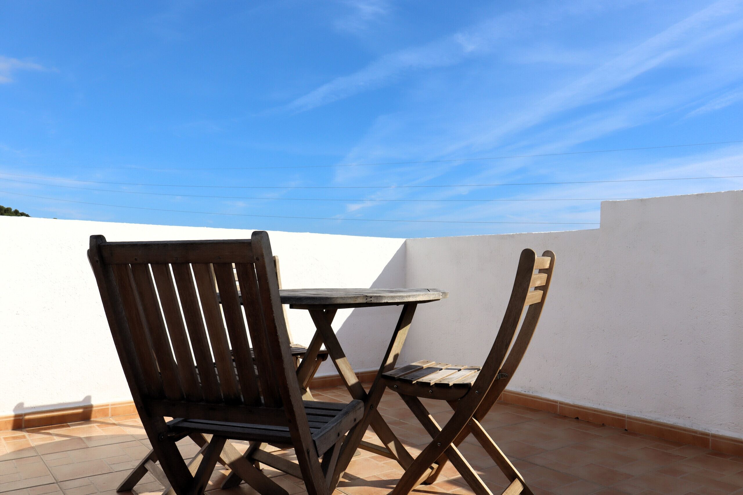 A small outdoor patio with a wooden table and two chairs, a white wall, and a clear blue sky.