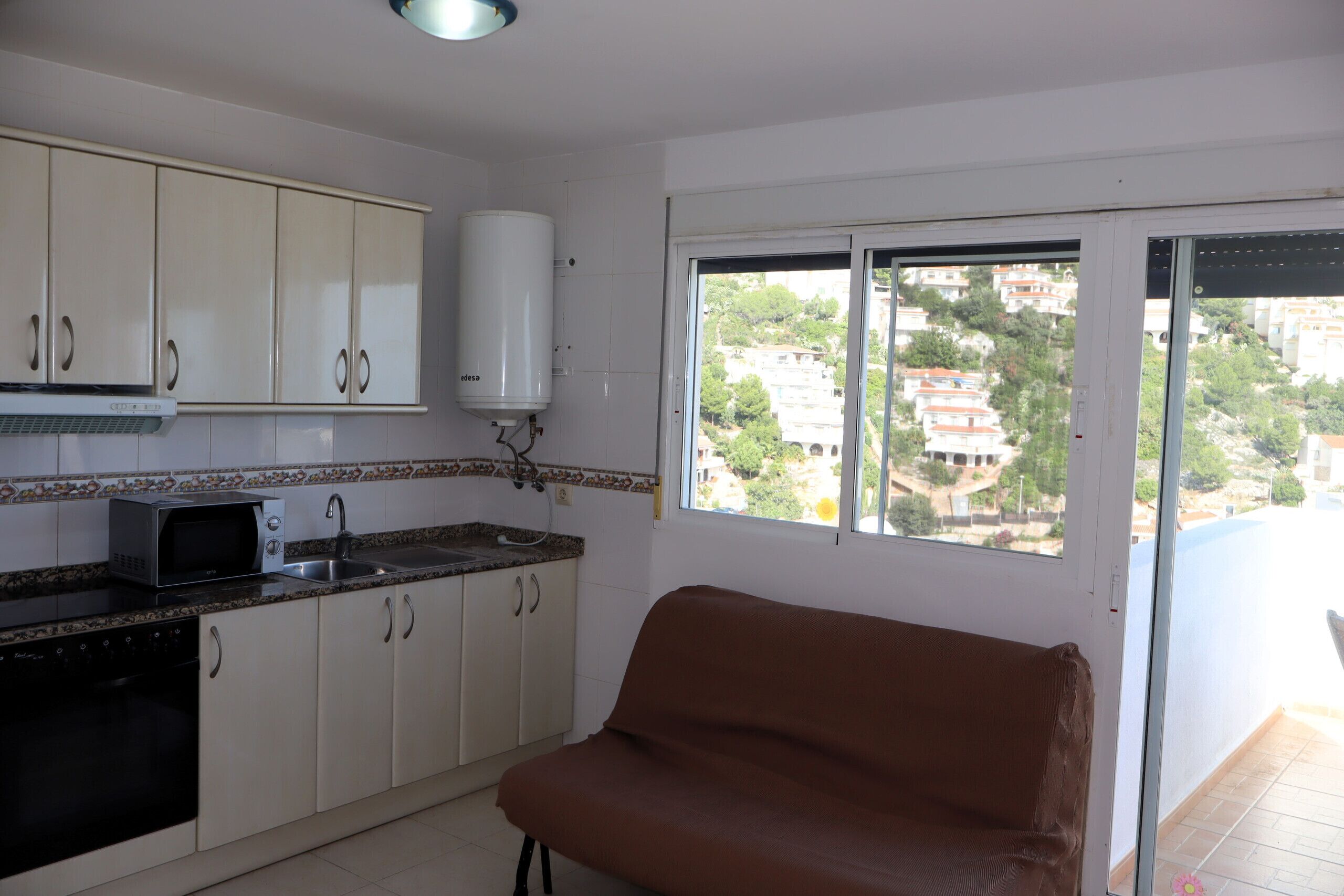 Una cocina con armarios blancos, microondas y un lavamanos. Hay una silla marrón cerca de una ventana con vistas a edificios.