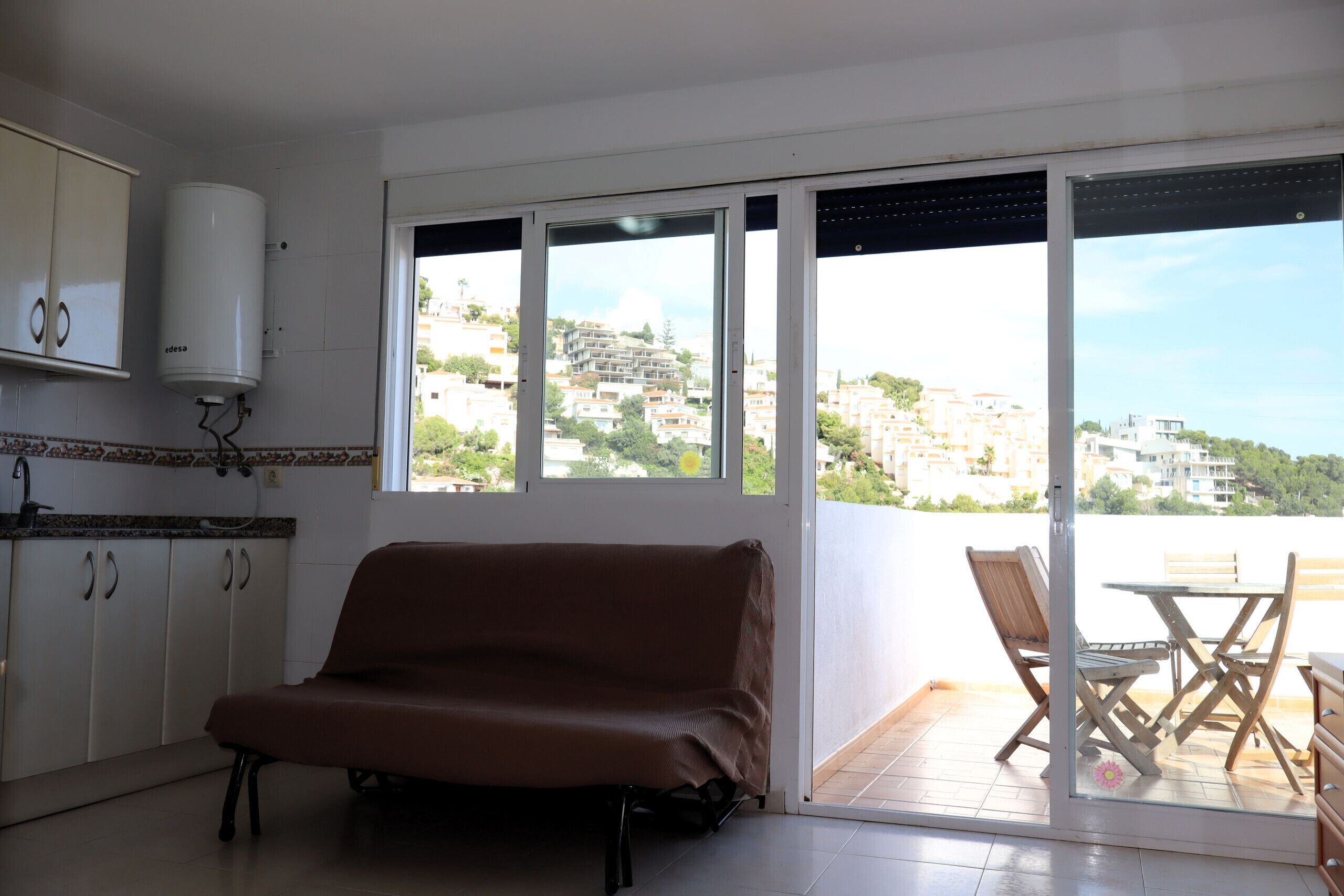 Habitación con sofá, mesa de comedor con sillas, zona de cocina y puerta corrediza de vidrio que da a un balcón con vistas a edificios.