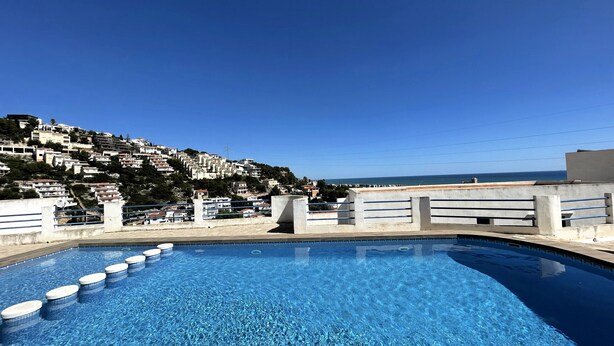En swimmingpool med klart, blått vatten, omgiven av en betongplattform och ett räcke. I bakgrunden syns en sluttning med byggnader och en klar himmel.