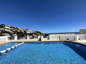 Une piscine à l’eau d’un bleu limpide, entourée d’une terrasse en béton et d’une rambarde. Au fond, on aperçoit une colline avec des bâtiments et un ciel dégagé.