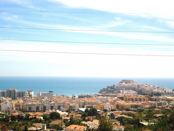 Un paysage urbain côtier avec une imposante forteresse perchée sur une colline, des bâtiments urbains denses et une vue dégagée sur la mer.
