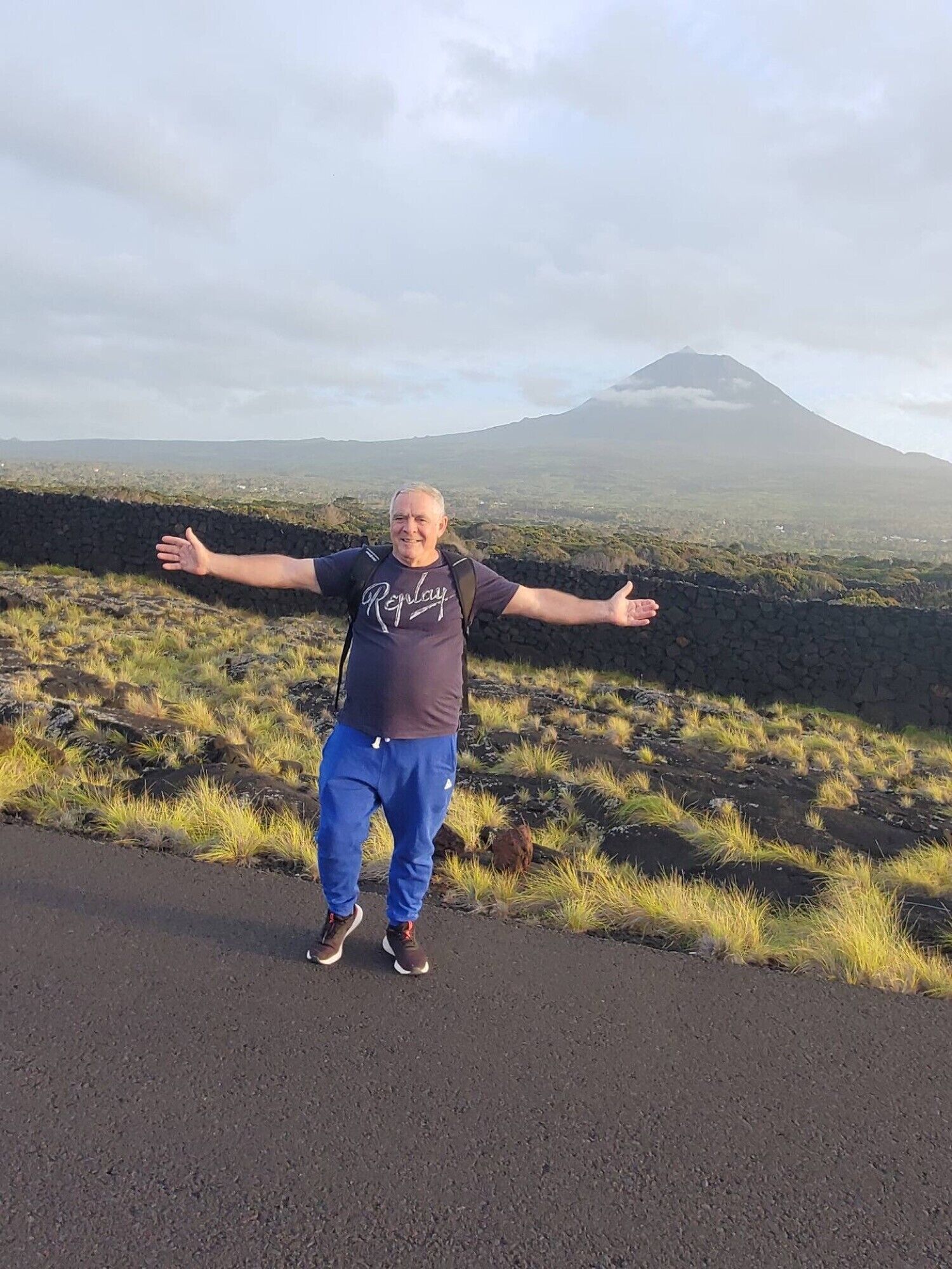 A person standing on a road with arms outstretched, facing a mountain in the background.