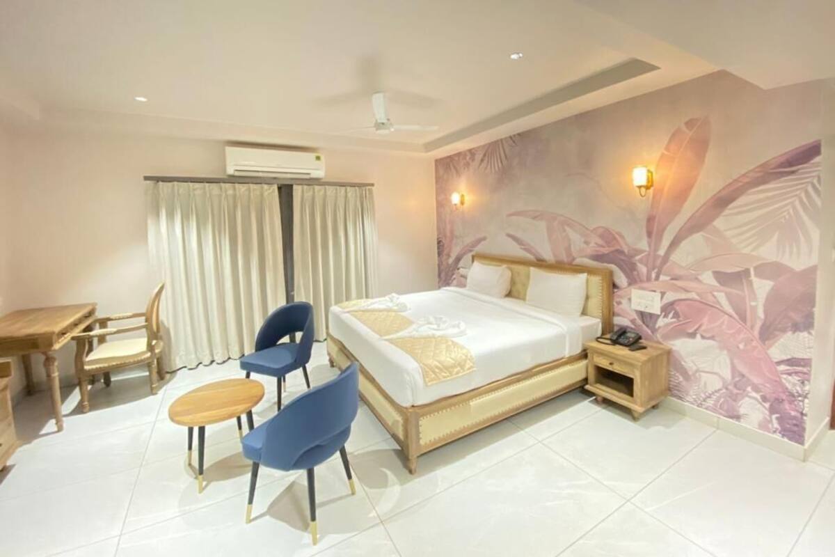 A hotel room with a bed, two chairs, a small table, an air conditioning unit, and a wall with a tropical mural.