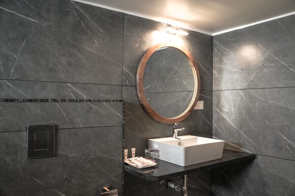 A modern bathroom with a marble wall, a round mirror, a white sink, and a black countertop. - Ariena by ABA Hotels (Morjim)