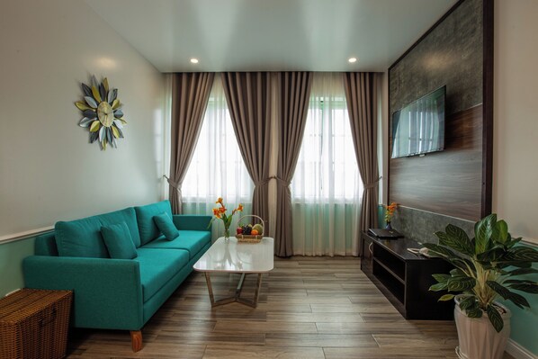 A modern living room with a teal sofa, a coffee table, a TV mounted on the wall, and a large window with curtains. - HARRY PHU QUOC HOTEL 3 (Phu Quoc)