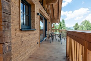 Una terraza de madera con una mesa y sillas, barandilla de madera y vistas a árboles y montañas.