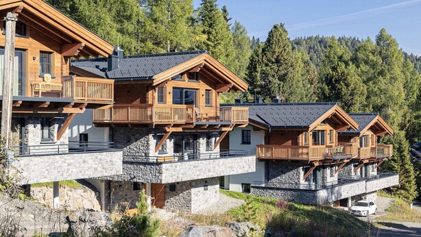 Un chalet en bois à plusieurs étages, doté de balcons, de fondations en pierre et d’une voiture garée devant.
