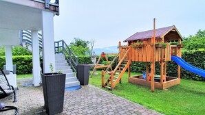 Una casita de juegos de madera con un tobogán, una escalera y un jardín con una maceta y muebles de exterior.