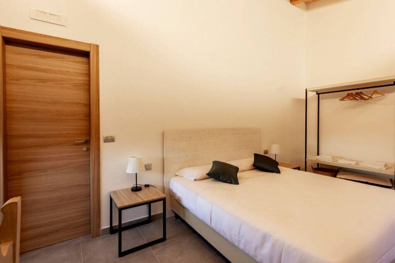 A bedroom with a wooden door, a small bedside table with a lamp, a bed with two pillows, and a mirror above a shelf with clothes.